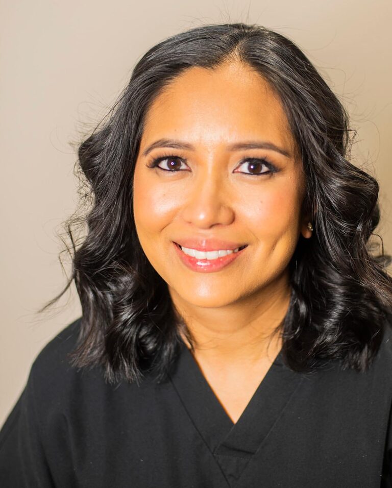 Portrait of a smiling woman with shoulder-length wavy black hair, wearing a black V-neck top. She is posed against a plain light background, suitable for professional or personal identification use.