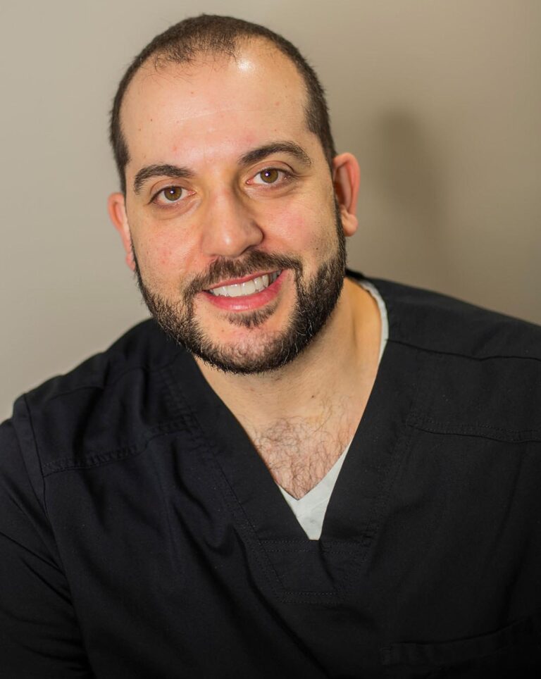 Portrait of a man with a trimmed beard and short hair, wearing dark medical scrubs and seated against a neutral background. His attire and setting suggest a healthcare professional, such as a doctor or nurse.
