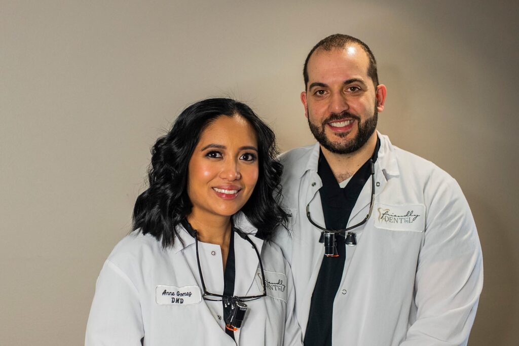 Two smiling dental professionals stand side by side in white lab coats with dental loupes around their necks. One coat reads “Anna ,DMD” and the other displays the “FRIENDLY DENTAL” logo, suggesting a clinical setting.