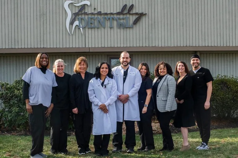 Group portrait of nine individuals standing in front of a building labeled “Friendly Dental.” Some wear medical scrubs or lab coats, suggesting a cohesive dental team posed outdoors on a well-maintained property.