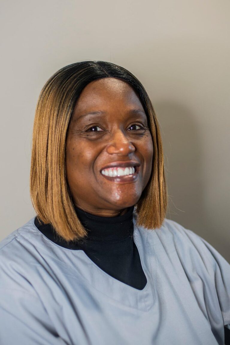 Portrait of a smiling woman with straight shoulder-length hair that transitions from dark roots to lighter brown. She wears a light gray top over a black undershirt, posed against a neutral background.