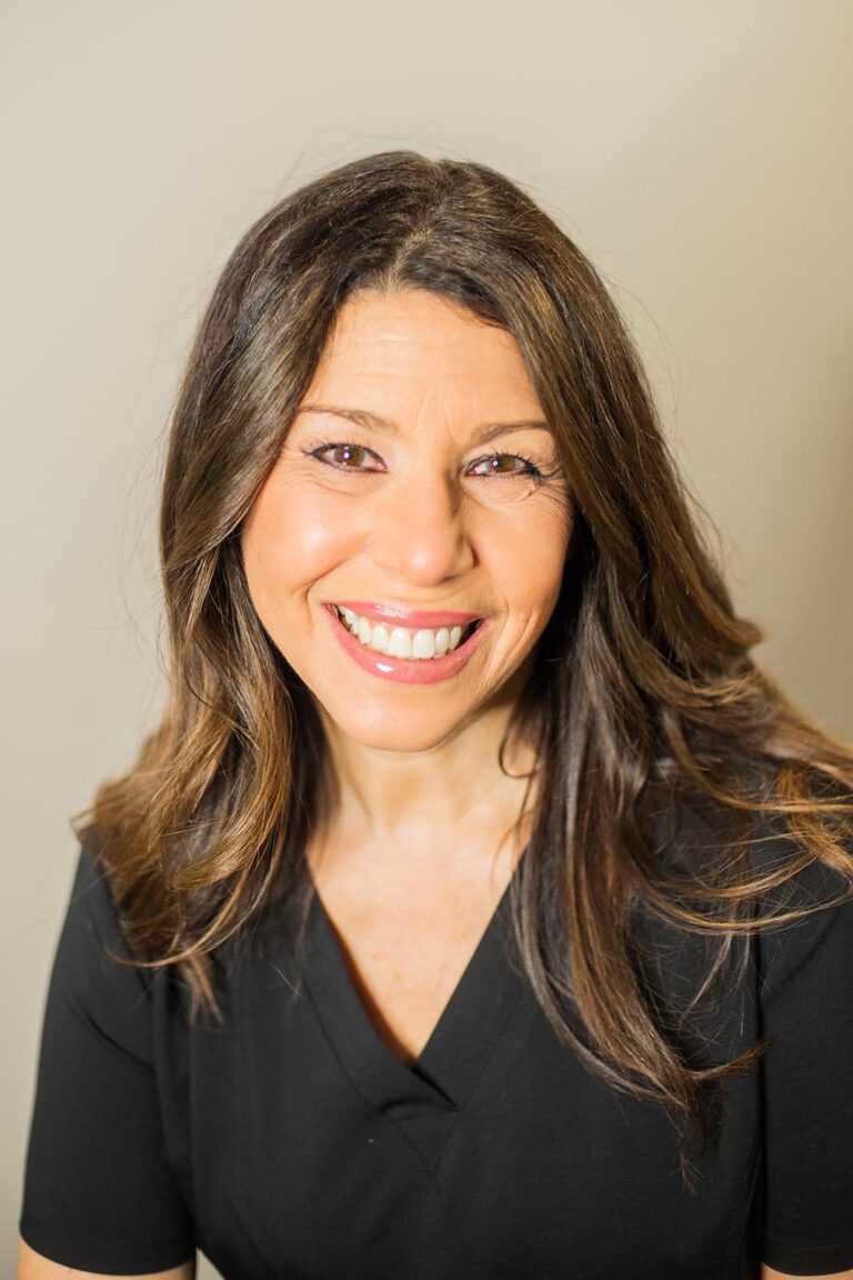 Portrait of a smiling woman with long, dark brown wavy hair, wearing a black top. She is posed against a plain light-colored wall with bright lighting that highlights her facial features.