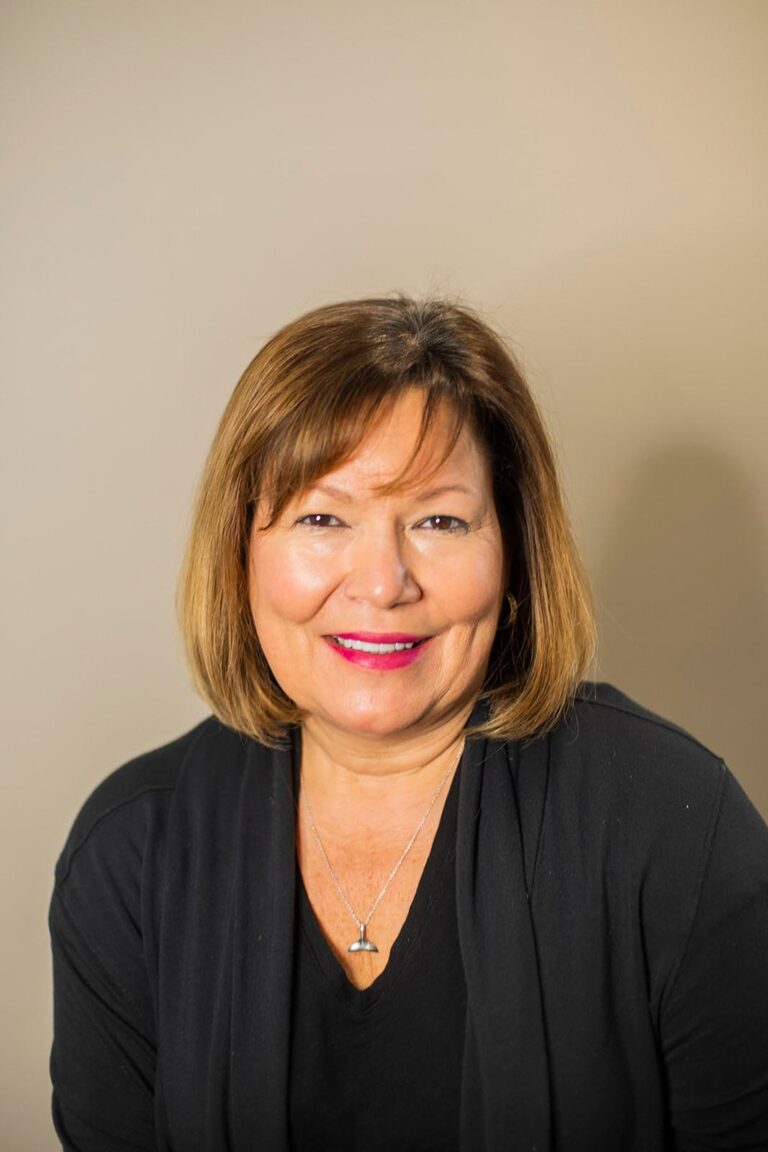 Portrait of a smiling woman with shoulder-length light brown hair, wearing a black top and a silver pendant necklace. She is posed against a plain beige background with bright lighting highlighting her face.