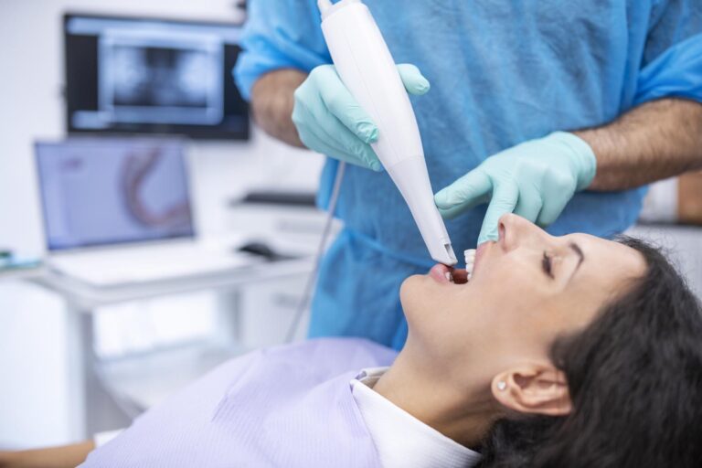 Dental procedure showing a gloved clinician using an intraoral scanner on a patient in a reclined chair. A monitor in the background displays dental images, highlighting the use of digital scanning technology.